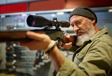 Bearded male hunter buying hunting rifle with optical sight in gun store. Weapon shop interior, ammo and ammunition assortment, firearms choice, shooting hobby